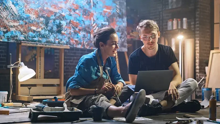 A woman holding a paintbrush near art supplies and man sit on the floor looking at a laptop computer screen. A large abstract painting hangs behind them.