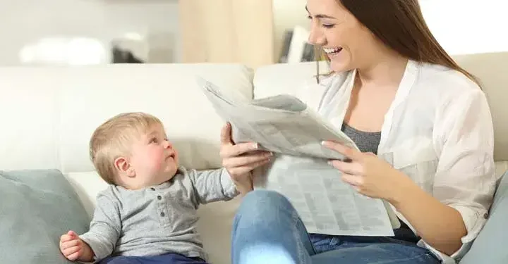 Mother reading a newspaper, while smiling at an infant sitting next to her on a couch