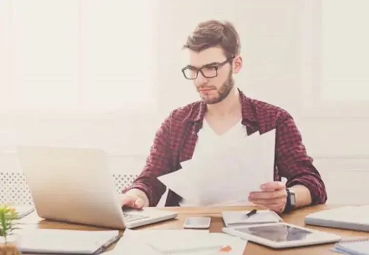 Man sitting at a cluttered desk holding papers and using his laptop