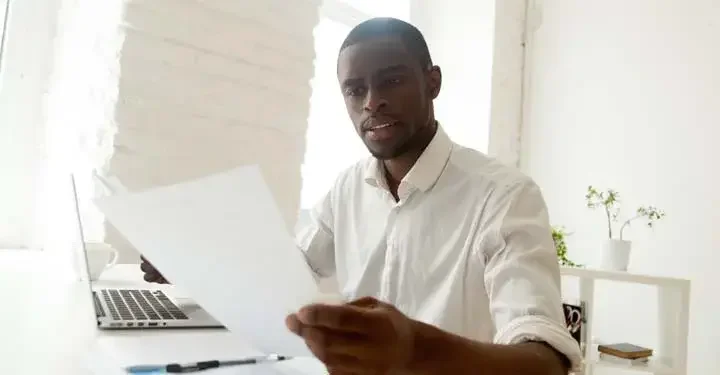 Man seriously looking at two sheets of paper above laptop and pen on white desk