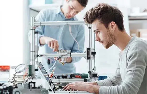 Person working on laptop in robotics lab while person in background holds machine