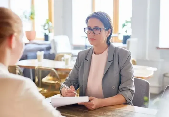 Woman in pink shirt and grey blazer and glasses signs document while gazing at woman across table