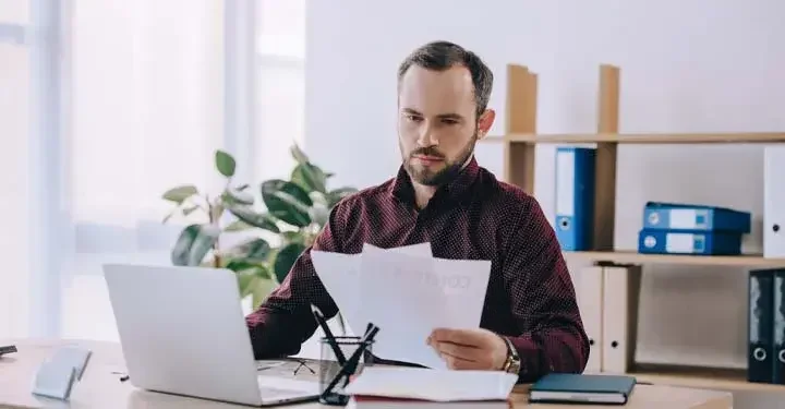 Man at desk in front of laptop and pencils in glass jar holding sheets of paper