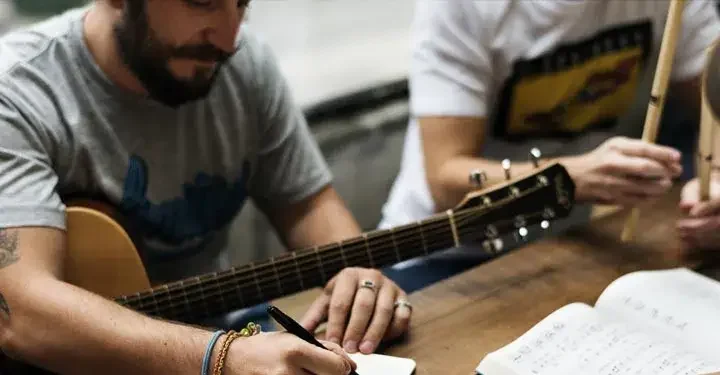 A man with a guitar writes music in a notebook, while a man holding drumsticks sits next to him