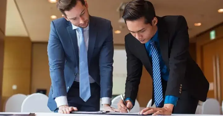 Two men in suits standing over a binder of papers while one signs a paper