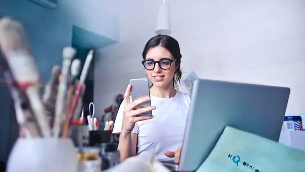 A woman checks her cell phone while seated at a desk with a computer and mug full of paintbrushes.