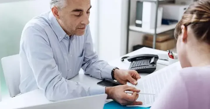 Man gesturing to sheet of paper with pen in hand while talking to another person next to landline phone