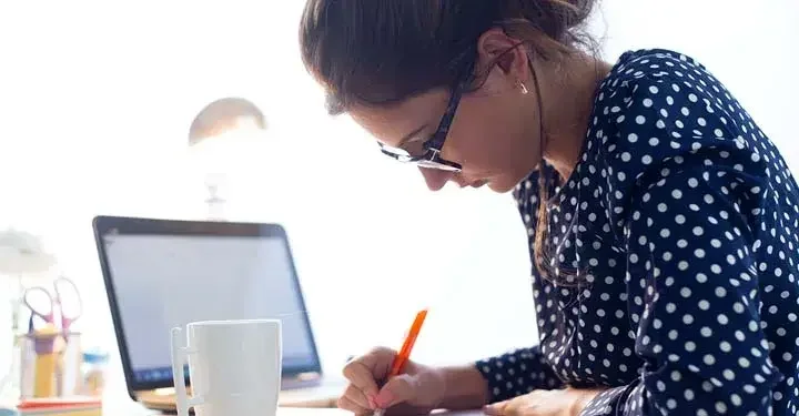 Woman holding pen and writing at a table with a cup of coffee and her laptop next to her