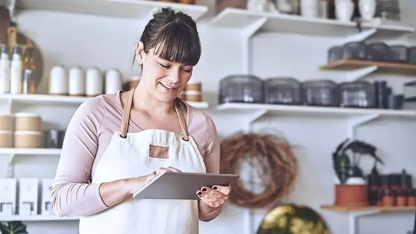 A woman in an apron stands in front of shelves of merchandise while working on her iPad.