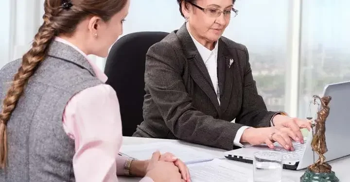 Older woman typing at laptop in suit while younger woman with blonde braid watches