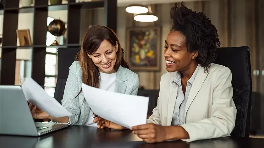 Two women, one with an open laptop, sit at a table and look over paperwork. Transforming your company's digital operations requires a strong strategy. 