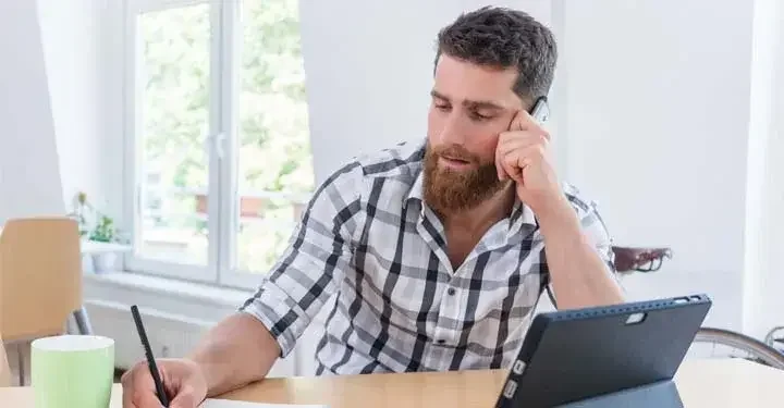 Man with an open tablet talking on the phone and writing on a piece of paper