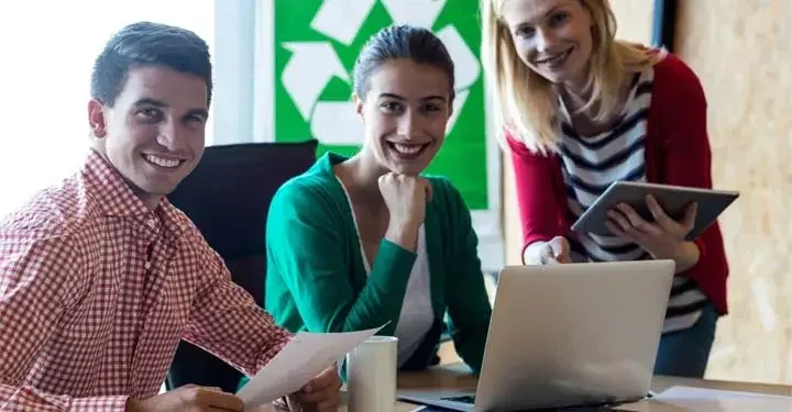 Three smiling coworkers with papers, a laptop, and a tablet