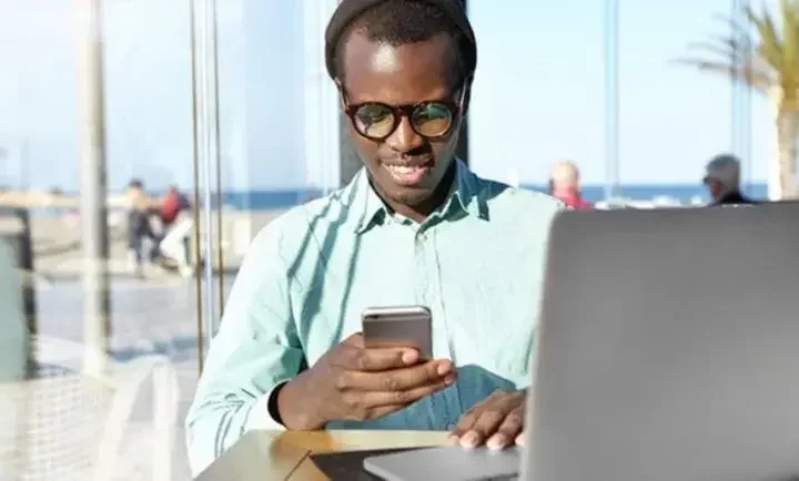 Man at a table next to a window overlooking the beach looking on his phone with his laptop open