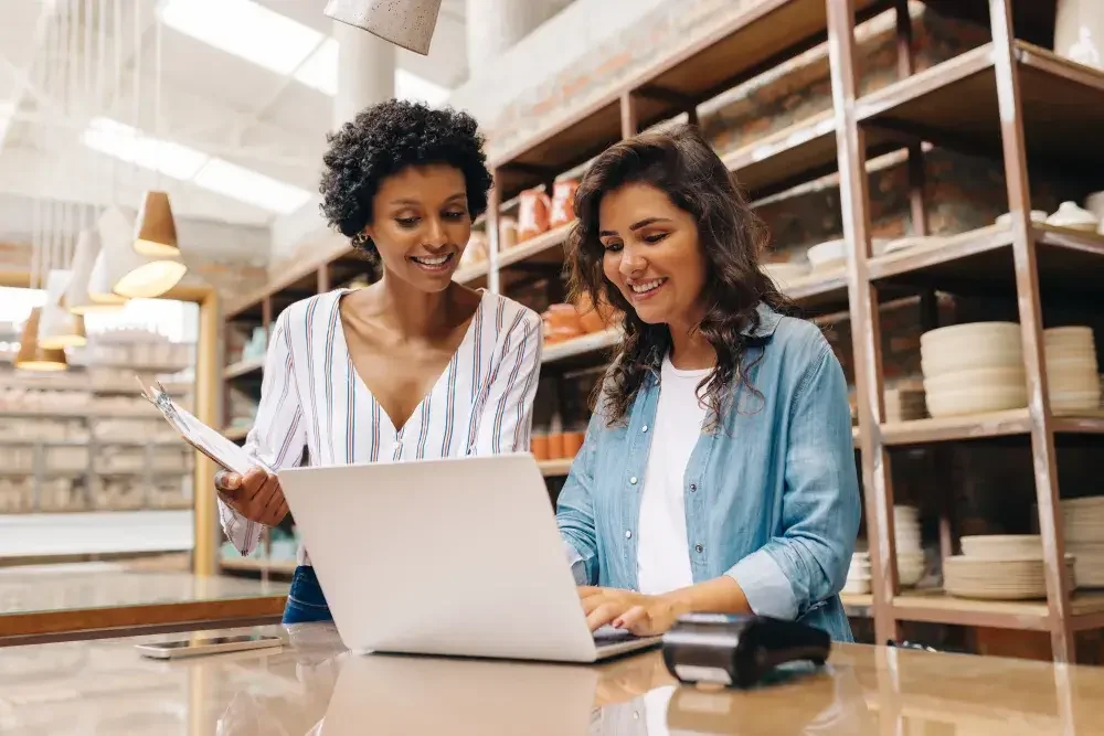 Two women stand behind the counter of a housewares store while looking at the screen of an open laptop to form their LLC..