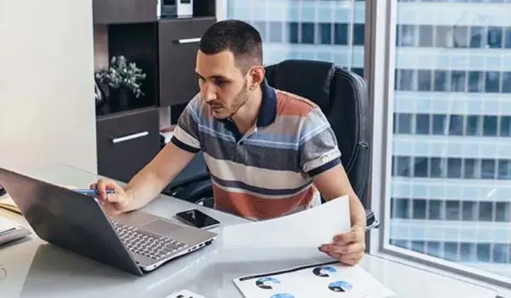 Man in office looking at computer holding a paper and pen with phone on desk