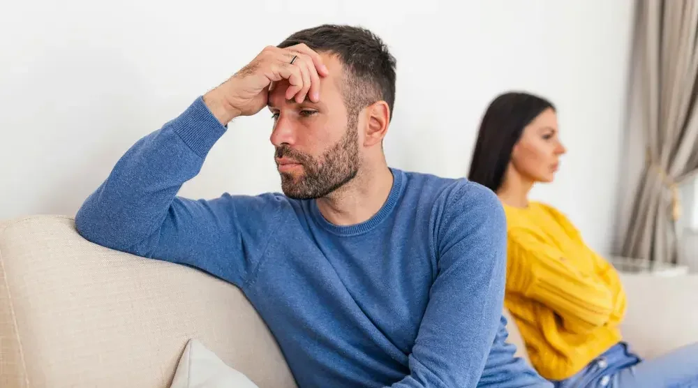 A man in the foreground sits on a couch with his back to his wife, who sits on the other end with her arms crossed. When dissolving a marriage, divorce is often easier than annulment. 