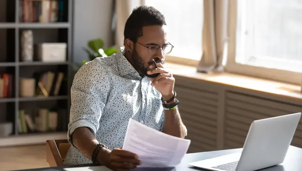 A man wearing glasses seated at a desk holds forms in one hand while viewing an open laptop.