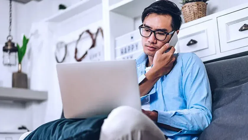 serious-man-listens-on-phone-looking-at-laptop wearing a blue shirt 