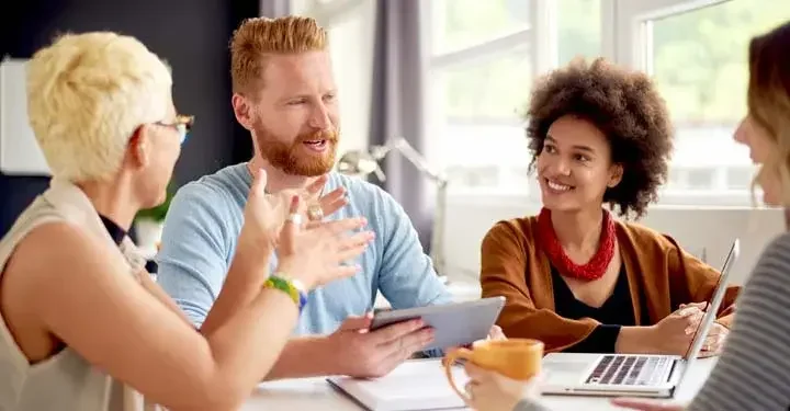 Group of four people sitting around table and smiling and talking with laptops and mugs