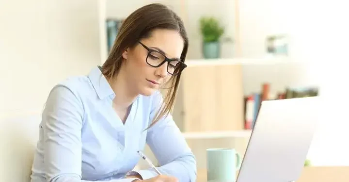 Businesswoman in glasses with long hair writing with ballpoint pen in front of laptop and coffee mug