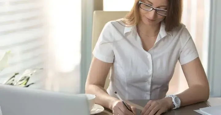 Woman writing with ballpoint pen next to coffee cup in front of laptop