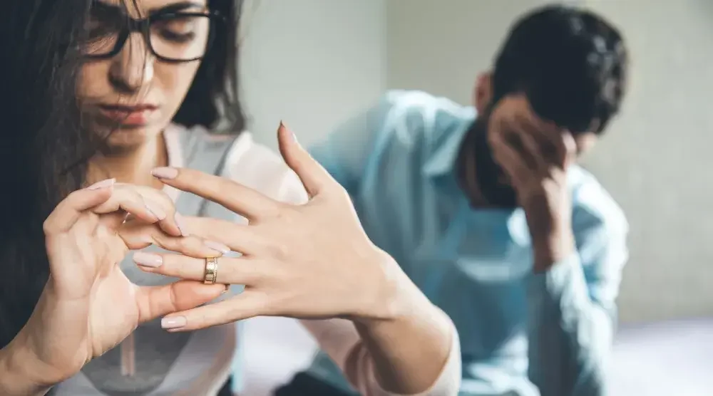 A woman removes her wedding ring while her husband sits behind her, holding his head in his hands.