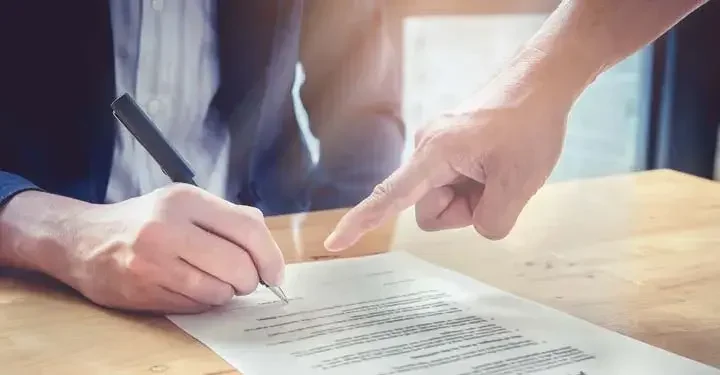 Person signing a document with another person's hand pointing at the paper