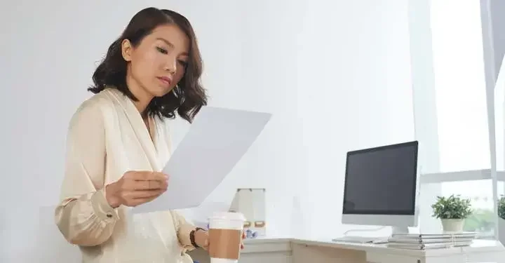 Woman with stylish bob looks at sheet of paper while holding disposable coffee cup with desk in background