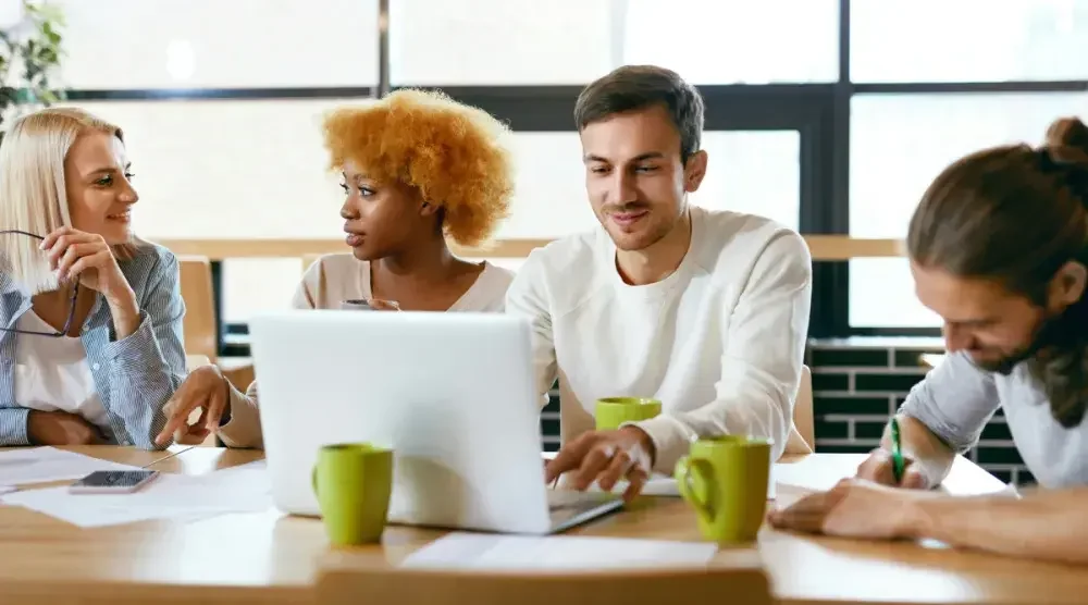 A group of four business partners in casual dress sit at a desk in an open, airy office and discuss their grant proposal.