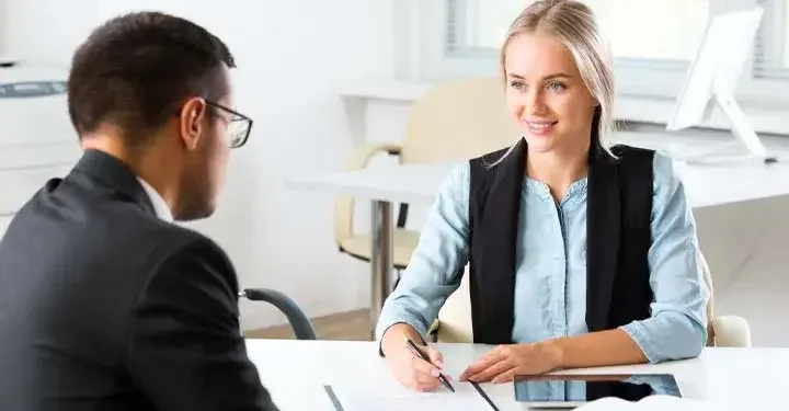 Smiling blonde woman holding pen over document with iPad on desk next to document while looking into eyes of man in blazer across from her