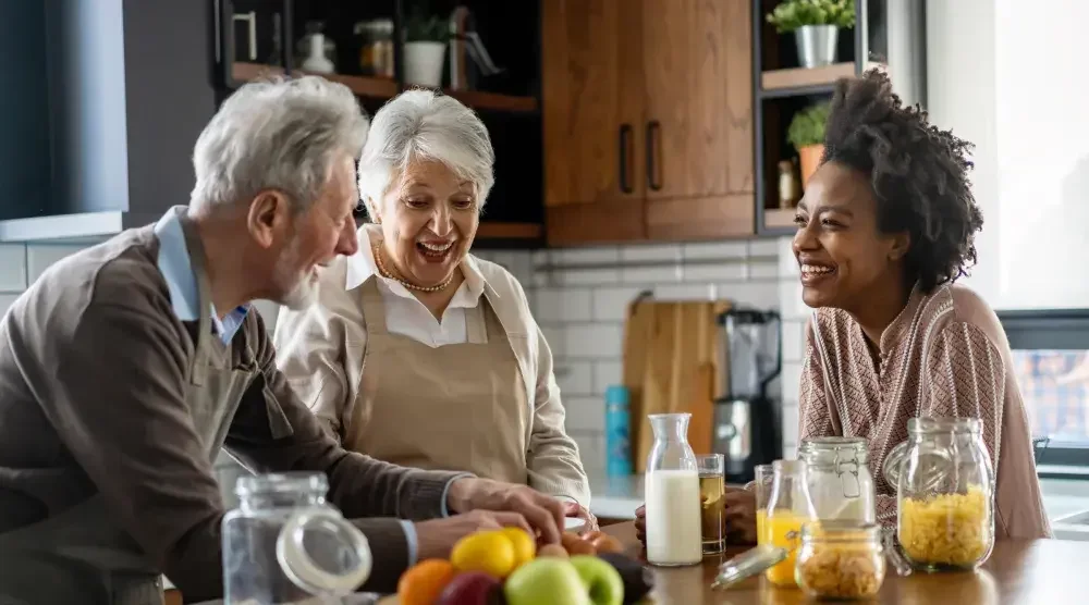 An elderly couple laughs with their adopted adult child in the kitchen.