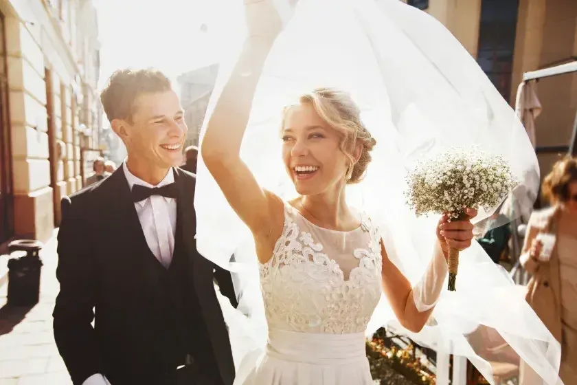 A newly married man and woman depart their wedding ceremony, with the bride holding her veil above her head. Some spouses prefer to keep their own names, while others are open to alternatives such as hyphenated last names.