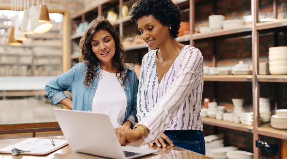 Two owners of a small housewares store stand behind a counter and look at their partnership agreement on a laptop computer.