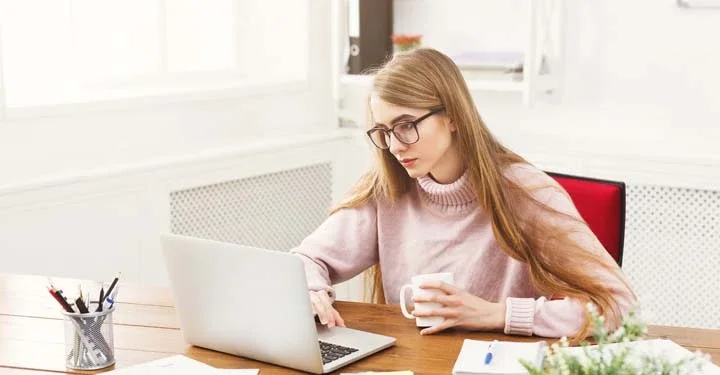 Woman wearing glasses holding a white mug using a laptop