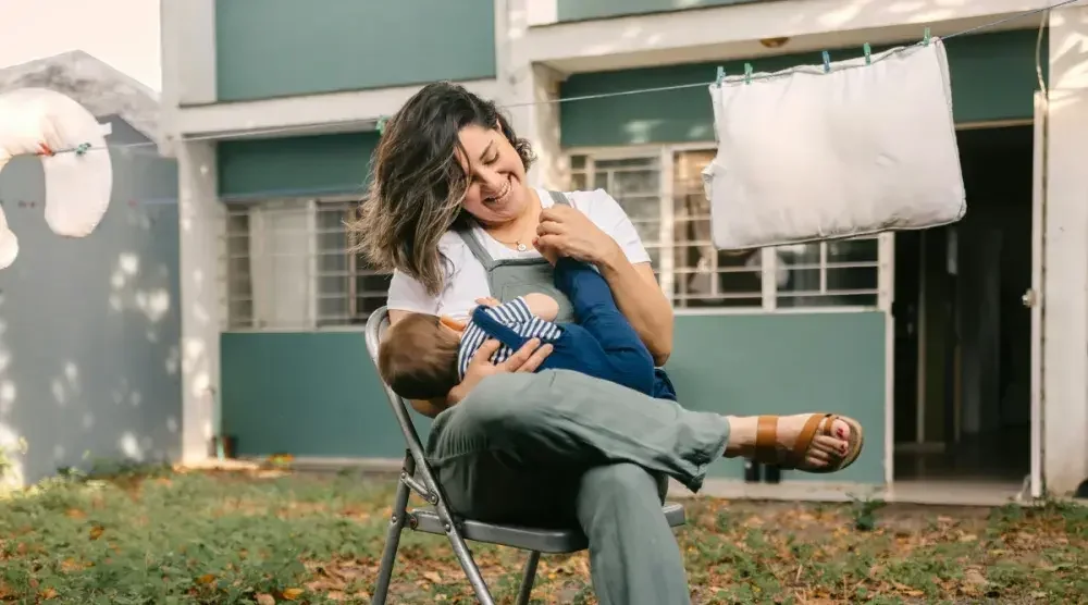 A woman in overalls sits on a folding chair in her yard while she plays with her baby in her lap.