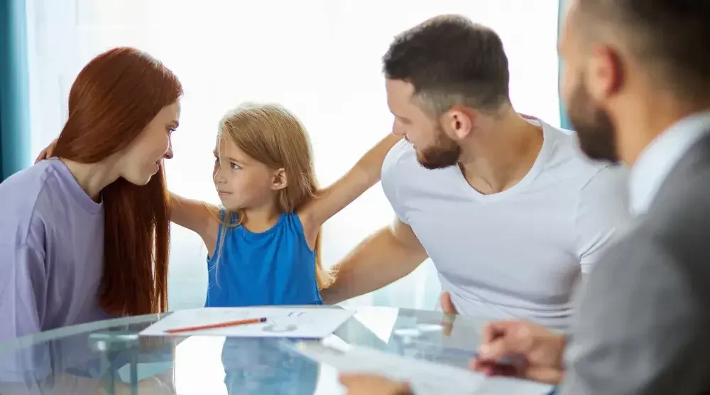 A young girl stands between her mother and father as they sit at a table with a divorce attorney