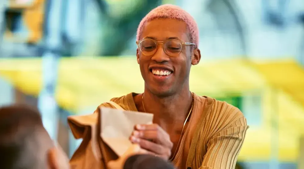 A black man in his 30s with pink hair hands over two paper bags to a customer at a farmer's market.