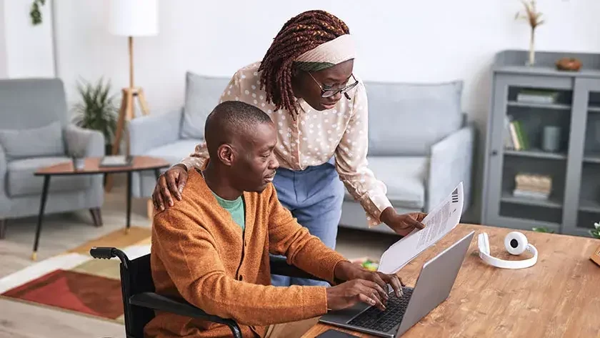 A woman leans over a man's shoulder as they look at a laptop displaying the website of their jointly owned business.