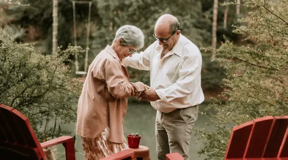 A man helps his wife stand from an adirondack chair in their back yard garden.