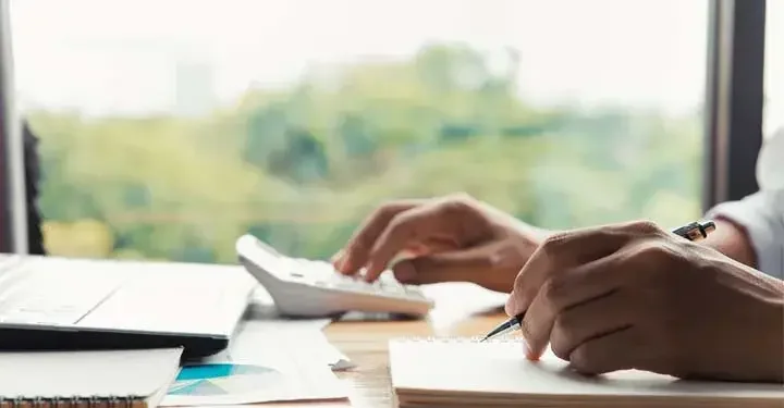 One hand holding a pen above a notebook while another hand uses a calculator in front of a computer and window