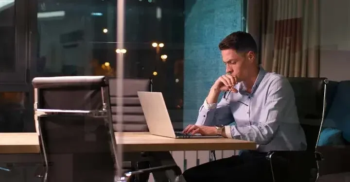 Concerned businessman sitting alone in front of laptop in sleek office at night in front of glass window showing city lights