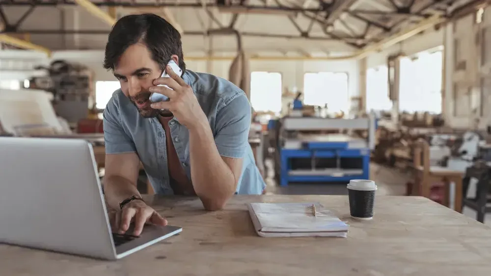 man talking on the phone looking at laptop in workshop