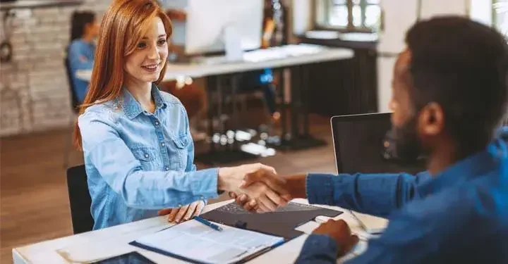 Man and woman shake hands at desk with papers and pen on it in office