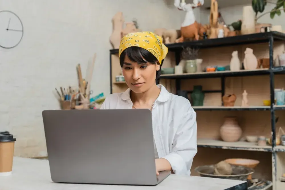 A woman wearing a bandana on her head sits at a counter in her pottery shop and uses her laptop computer. 