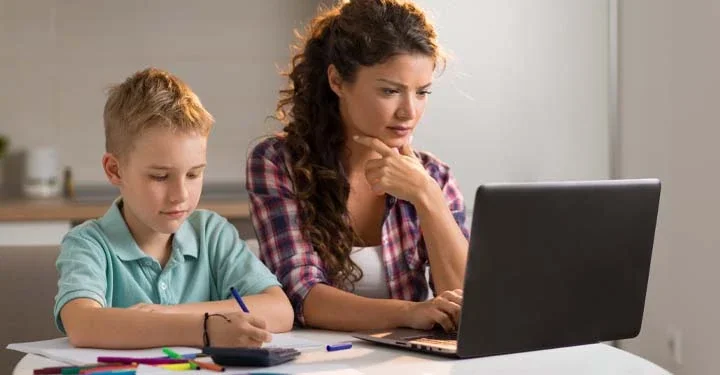 Little boy in pale blue polo coloring in a book while sitting next to a woman, who is using a laptop