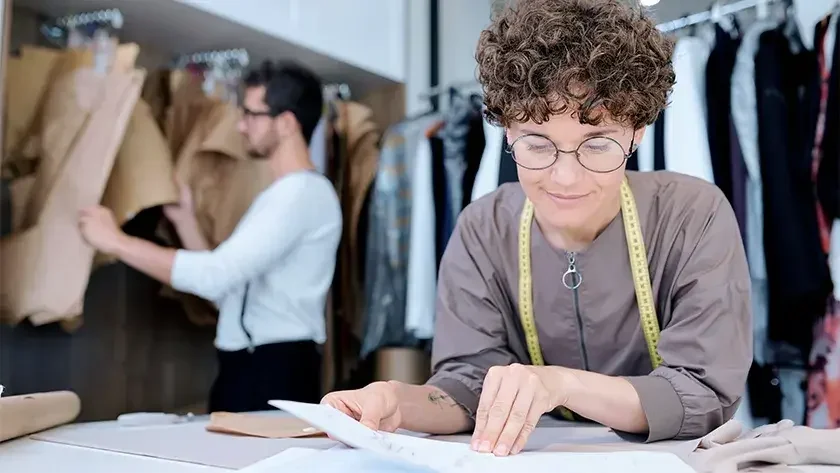 A woman with a tape measure draped around her shoulders sits at a desk and looks over LLC formation documents. Behind her, an employee goes through racks of clothing.