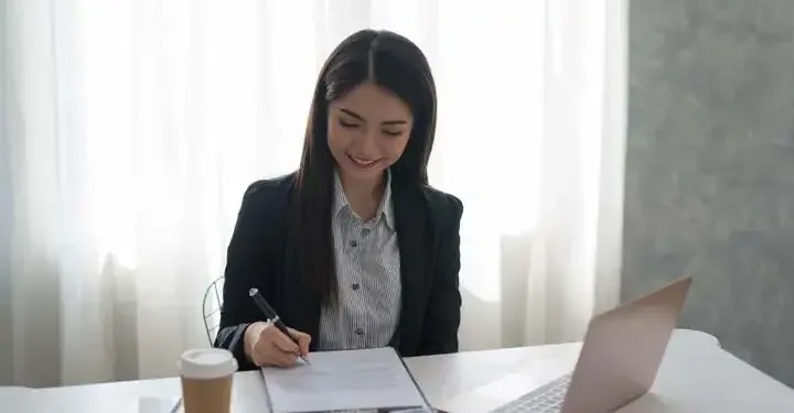 Woman smiling while signing form in front of laptop and coffee cup