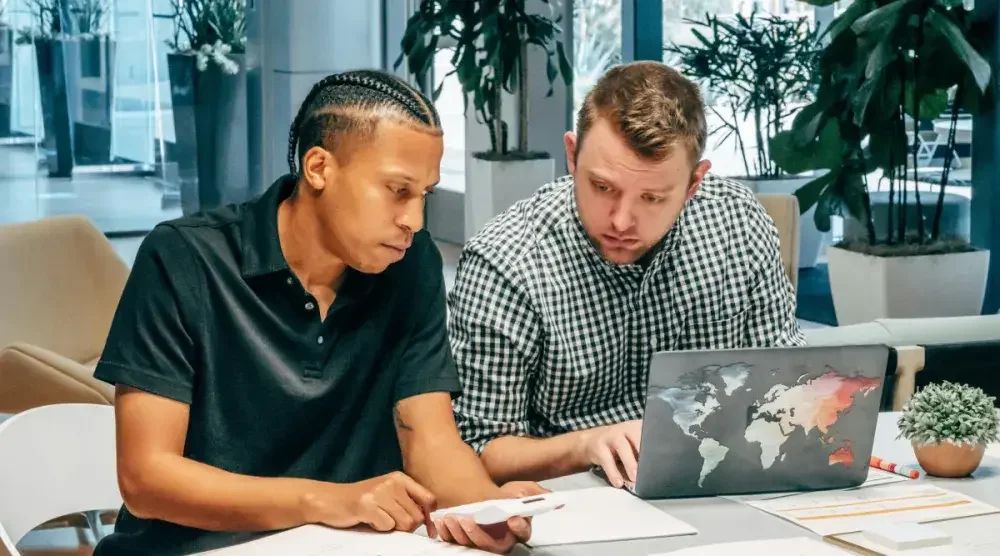 A black man and a white man sit at a desk together and review the results of a business search.