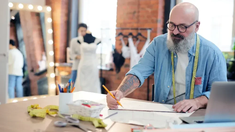 A tailor sits at a counter making a sketch as an employee does a fitting behind him.